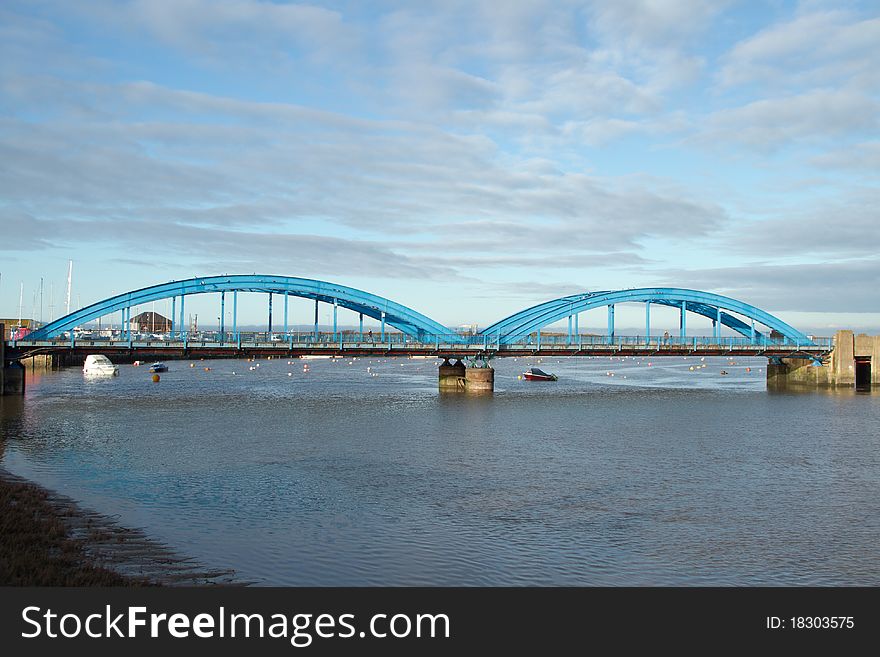 A double span blue painted metal bridge with concrete supports crossing a river with a blue cloudy sky. A double span blue painted metal bridge with concrete supports crossing a river with a blue cloudy sky.