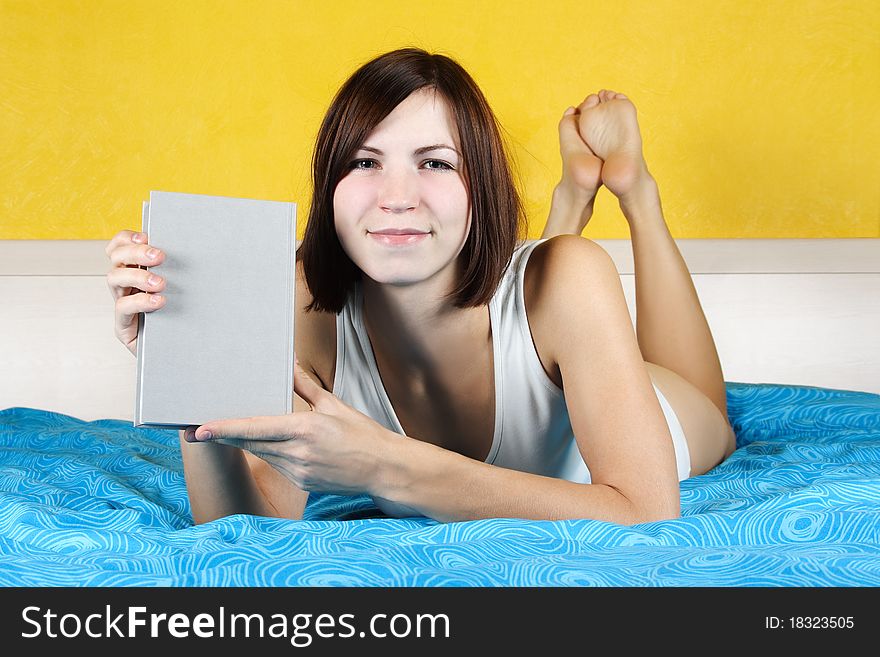 Young woman in white undergarment lying in bed on belly and showing book, bright interior. Young woman in white undergarment lying in bed on belly and showing book, bright interior