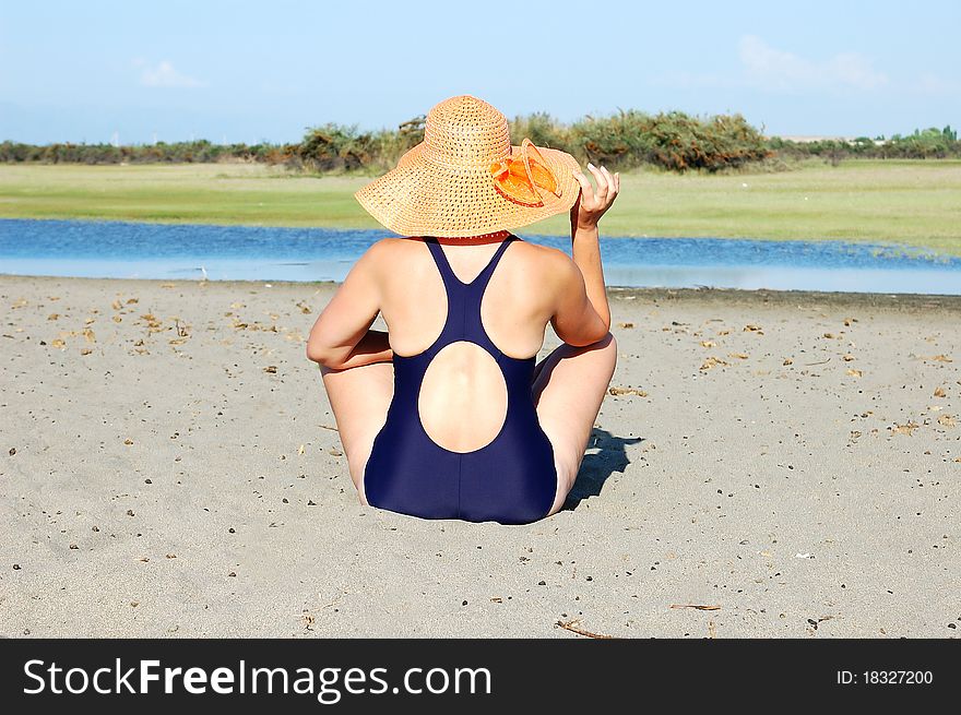 Woman in dark blue bikini sits on hot sand in a yellow hat to river bank