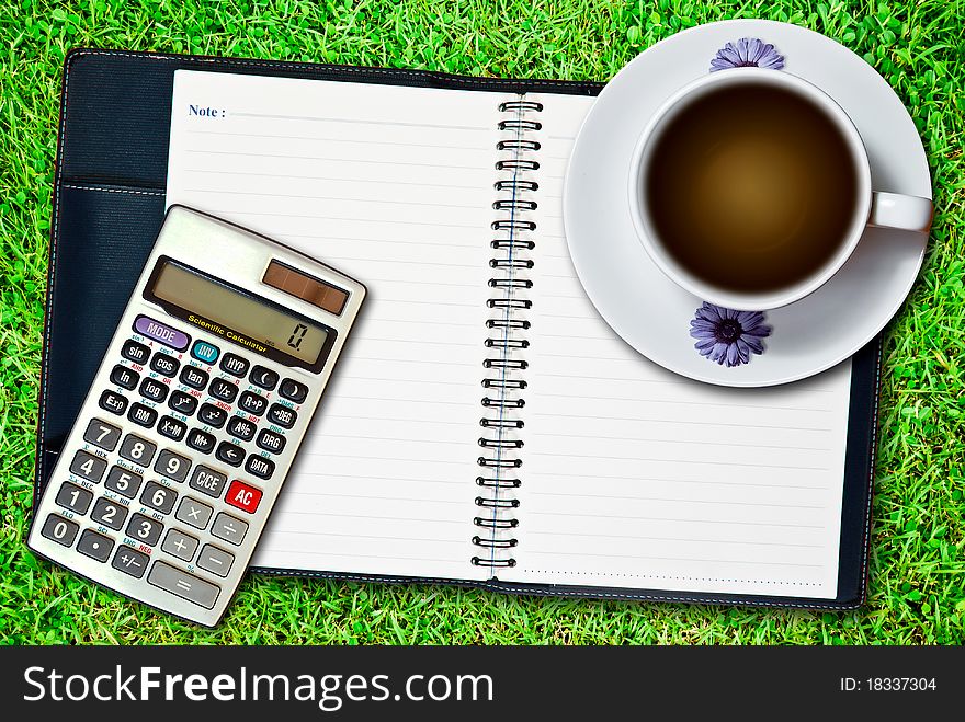 White cup of coffee and blank notebook with calculator on fresh green grass background. White cup of coffee and blank notebook with calculator on fresh green grass background