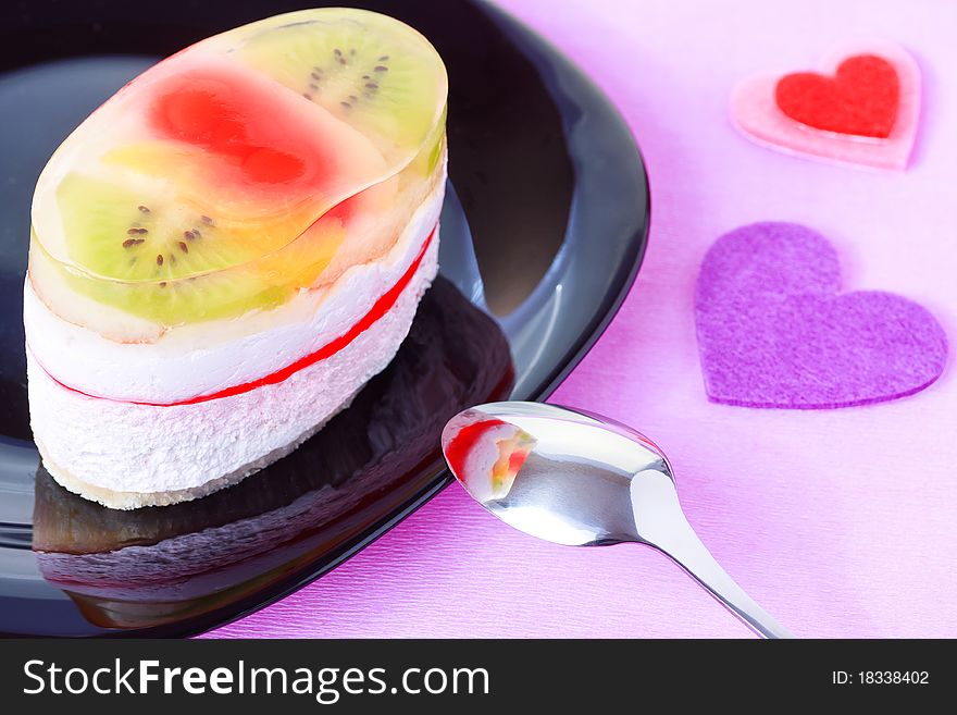 Closeup of cake with jelly and fruits on a black plate with Valentine`s day heart shaped decorations on a purple background. Closeup of cake with jelly and fruits on a black plate with Valentine`s day heart shaped decorations on a purple background