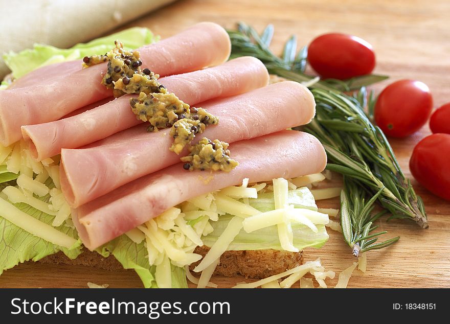 Tasty open sandwich with green lettuce, grated cheese, smoked ham and wholegrain mustard on wholewheat bread with rosemary and tomatoes on chopping board. Tasty open sandwich with green lettuce, grated cheese, smoked ham and wholegrain mustard on wholewheat bread with rosemary and tomatoes on chopping board