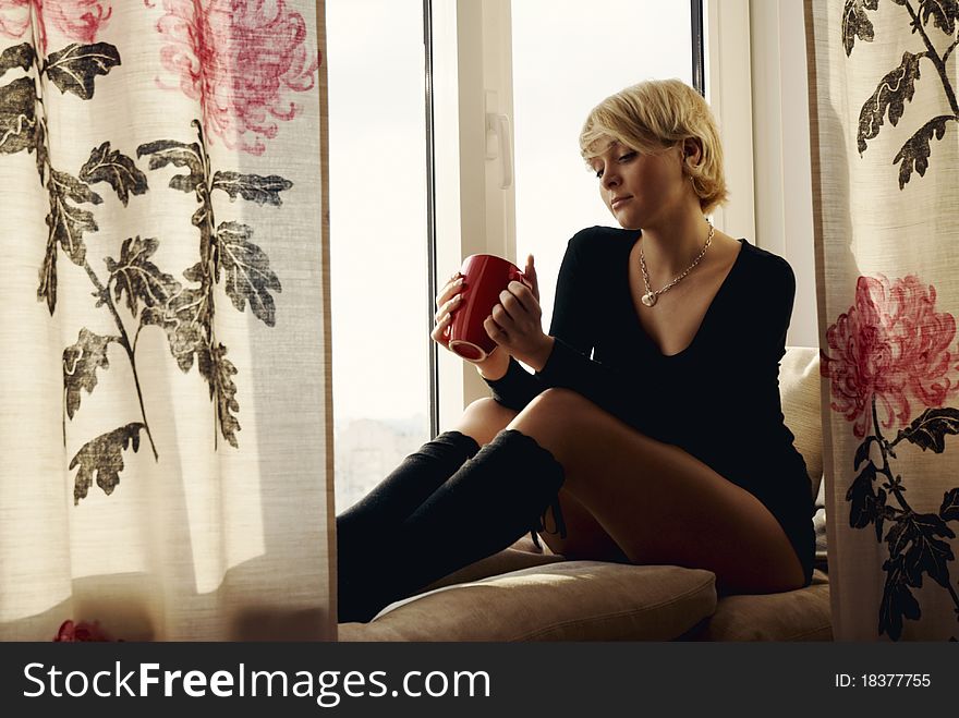 Portrait of charming girl sitting on windowsill and looking through window in the dusk