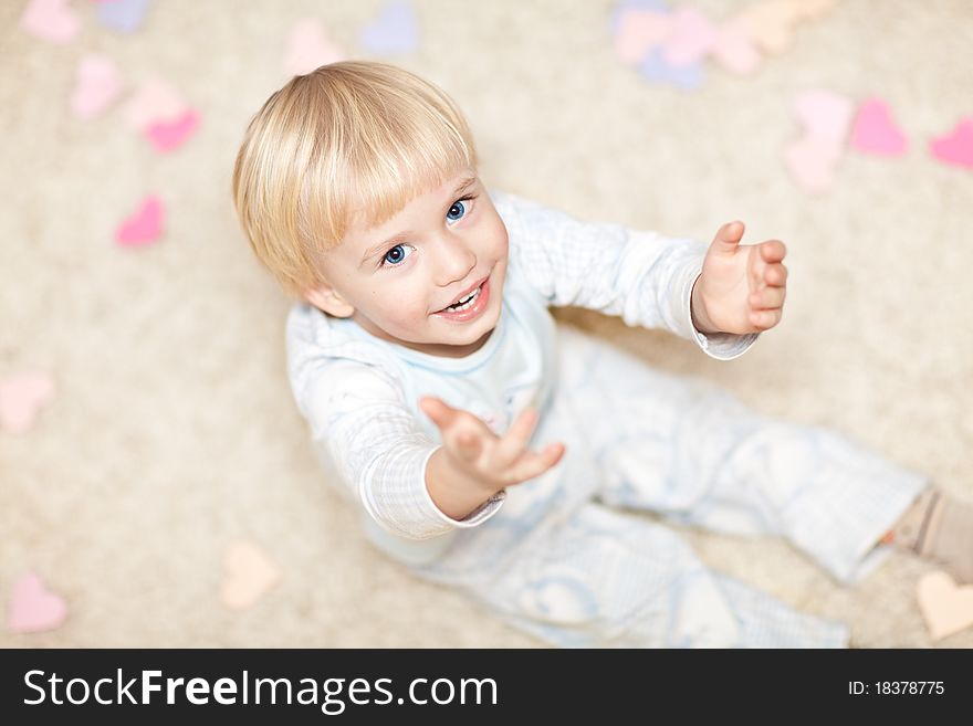 Sweet little boy sitting on the floor in pajamas and stretching his hands. There are little heart shapes around him.