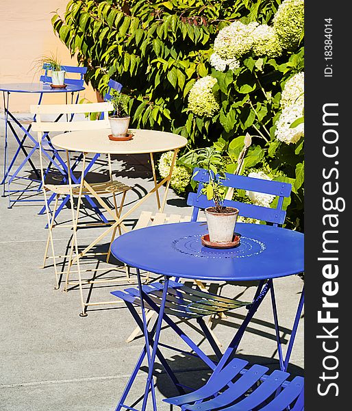 Row of colorful bistro tables outside at a sidewalk cafe. Flowerpot on each table and shrub behind. Nobody present. Row of colorful bistro tables outside at a sidewalk cafe. Flowerpot on each table and shrub behind. Nobody present.