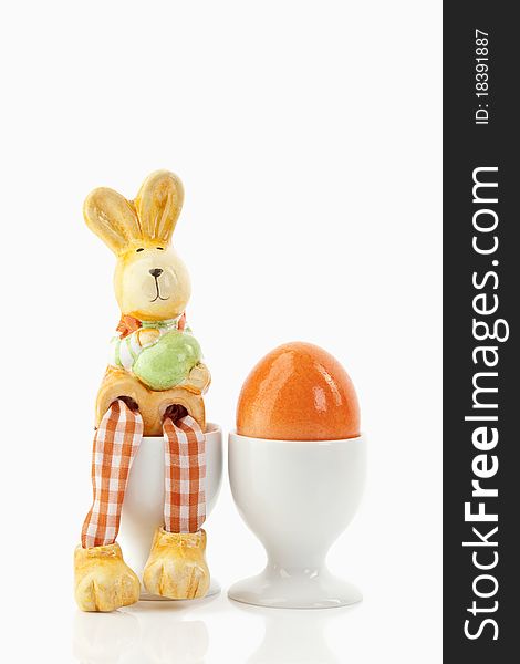 Easter bunny sitting on an egg-cup close to an orange egg in an egg-cup on a white background