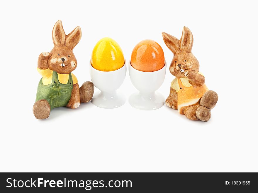 Two easter bunnies sitting close to orange and yellow eggs in egg-cups on a white background. Two easter bunnies sitting close to orange and yellow eggs in egg-cups on a white background