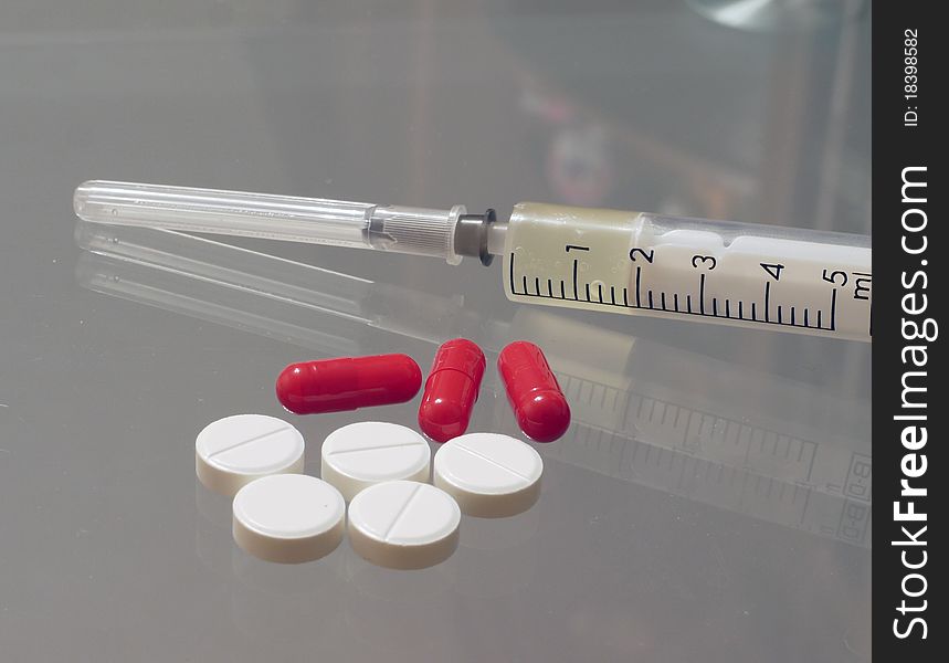 Five white round tablets, three red pills and one syringe lying on a glass table close up. Five white round tablets, three red pills and one syringe lying on a glass table close up