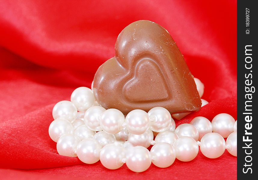 Chocolate heart resting on a string of pearls with red satin in the background in a vertical format. Could be used for Valentine, anniversary or romantic. Chocolate heart resting on a string of pearls with red satin in the background in a vertical format. Could be used for Valentine, anniversary or romantic