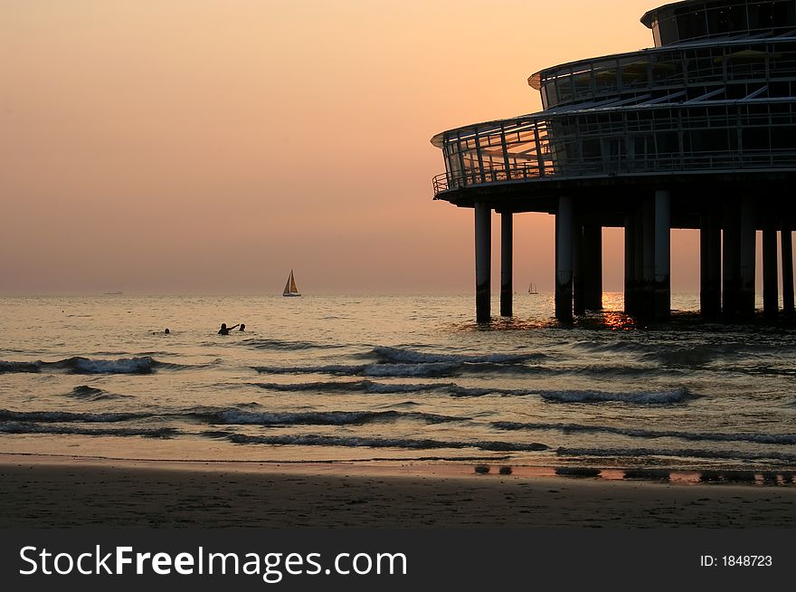 Sea with swimmers in the sunset, with a pier restaurant and a sailing boat on the horizon. Sea with swimmers in the sunset, with a pier restaurant and a sailing boat on the horizon