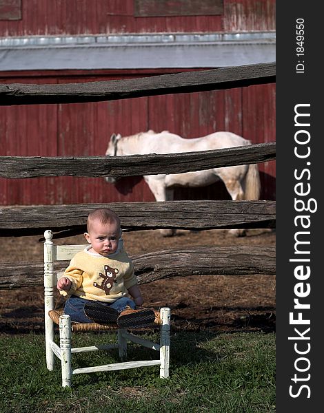 Image of baby boy sitting on a chair in front of a barn building, a horse in the background. Image of baby boy sitting on a chair in front of a barn building, a horse in the background