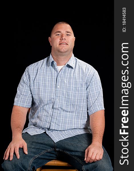 A young man sitting down on a wooden chair.  He has an odd look on his face.  He is wearing a blue shirt and there is a black background. A young man sitting down on a wooden chair.  He has an odd look on his face.  He is wearing a blue shirt and there is a black background.