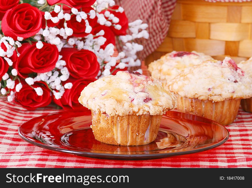 Raspberry muffin on a red glass plate with more muffins, roses, and part of a basket in the background all on a red checked tablecloth