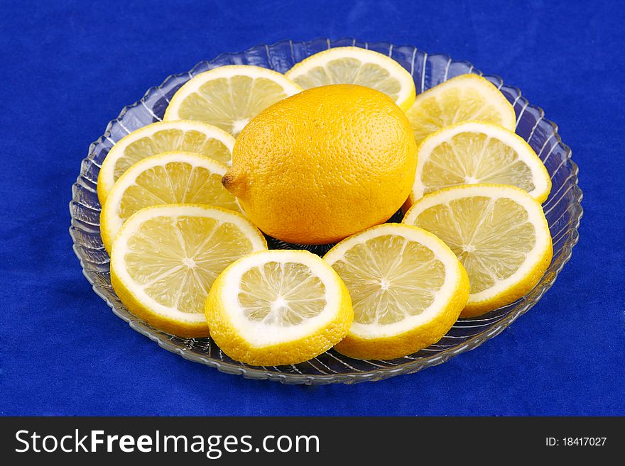 Whole lemon surrounded by lemon slices on a clear glass palte on a bright blue background. Whole lemon surrounded by lemon slices on a clear glass palte on a bright blue background