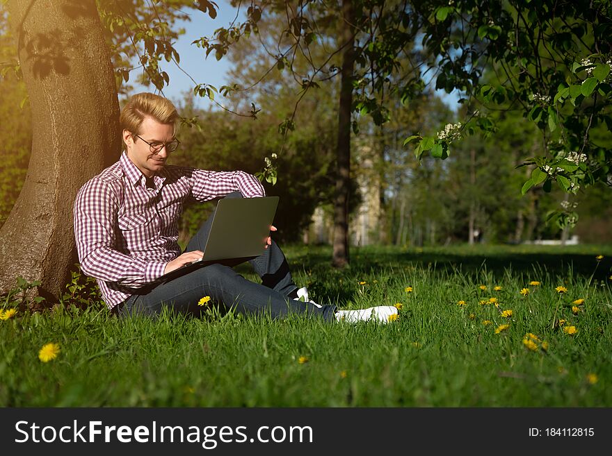 A Young Caucasian Man Sits Under A Tree, Typing On A Laptop. Freelancer Working In The Park On The Grass