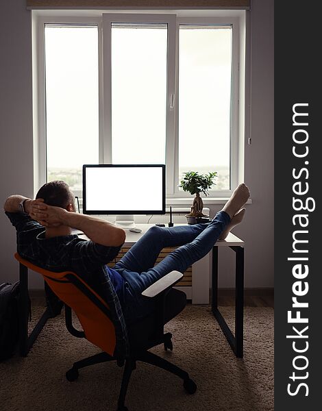 Man relaxing while working remotely from home holding legs on the table looking at the computer stretching hands behind his head. Man relaxing while working remotely from home holding legs on the table looking at the computer stretching hands behind his head