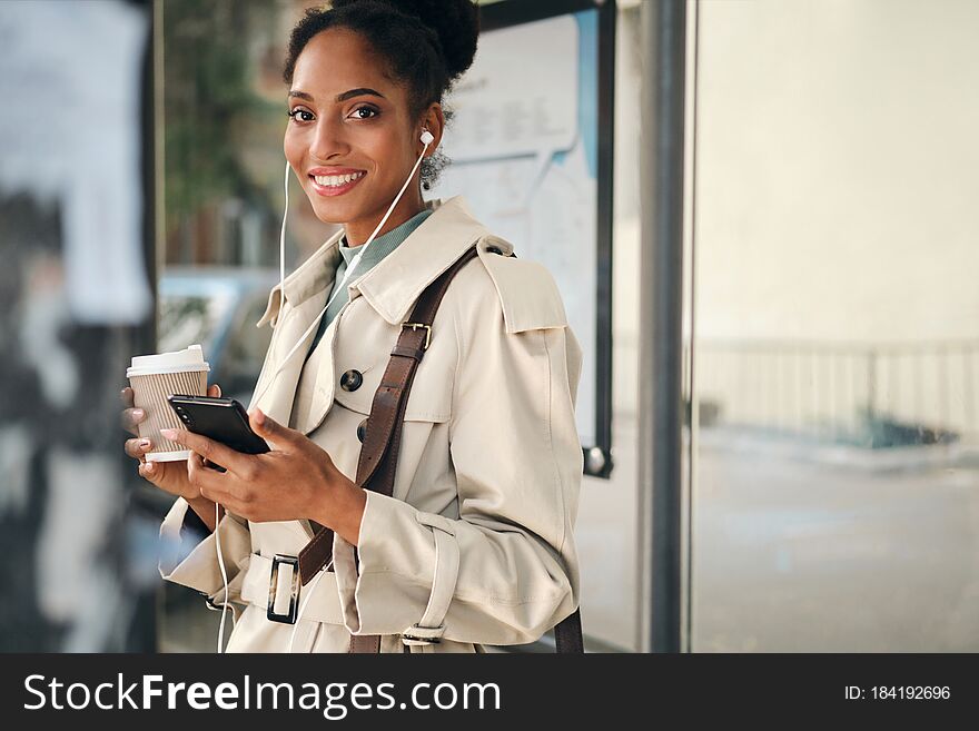 Beautiful casual African American girl in stylish trench coat with coffee to go and cellphone joyfully looking in camera at bus stop