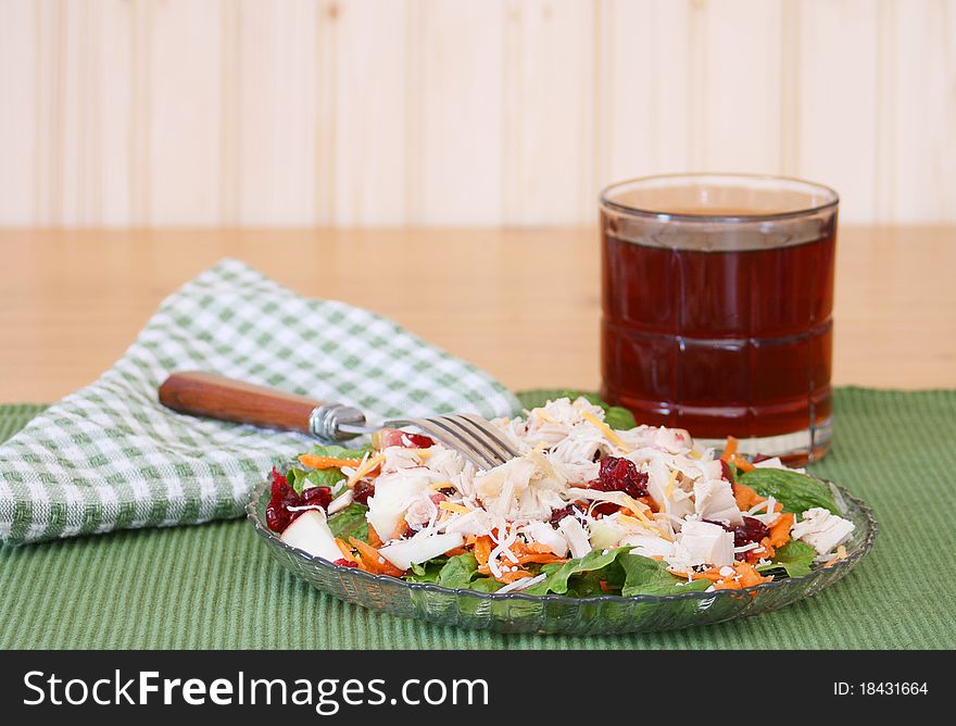 Salad with meat, vegetables and fruit on a glass plate with iced tea in the background on a green place mat with a green checked napkin and fork. Salad with meat, vegetables and fruit on a glass plate with iced tea in the background on a green place mat with a green checked napkin and fork