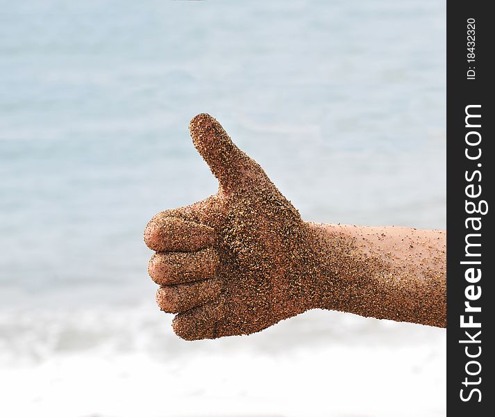 Beach fun with a hand with sand giving a thumbs up, with the ocean beach waves in the background.