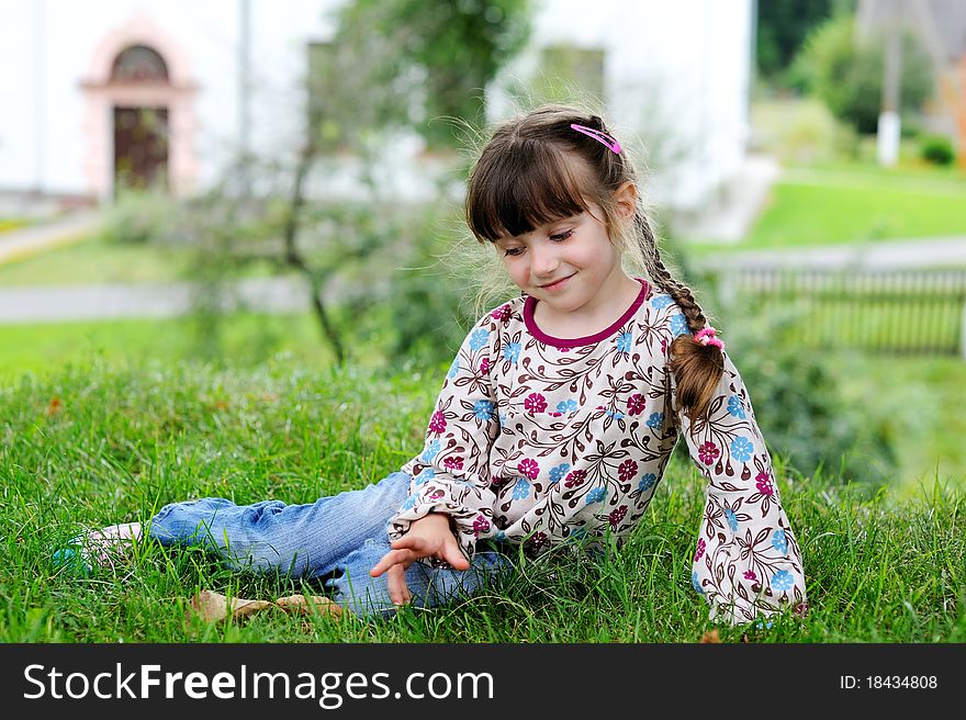Adorable little girl in colorful clothes sits on grass in front of old style house. Adorable little girl in colorful clothes sits on grass in front of old style house