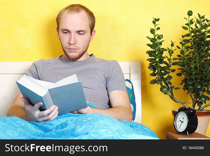 Young man lying in bed and reading book, table with plant and clock, bright interior