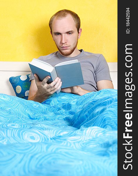 Young man lying in bed and reading book, bright interior