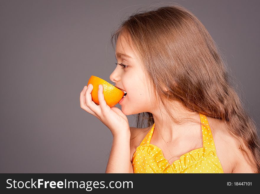Beautiful young cheerful girl has turned in profile and bites orange, against gray background