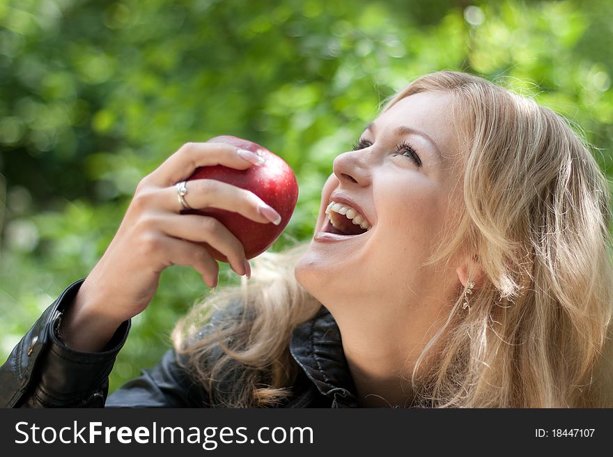 Beautiful young cheerful woman has turned in profile and bites red big apple, against green garden.