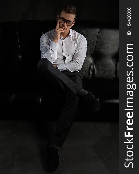 A handsome stylish confident bearded businessman in a white shirt and glasses is sitting on a black sofa in the office. A handsome stylish confident bearded businessman in a white shirt and glasses is sitting on a black sofa in the office.
