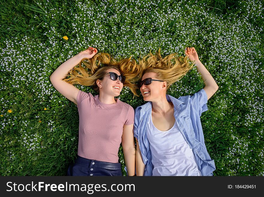Two Young Happy Women In A Flowery Meadow On Sunny Day, Top View, Close-up. Cheerful Female Friends Lying On The Lawn, Looking At