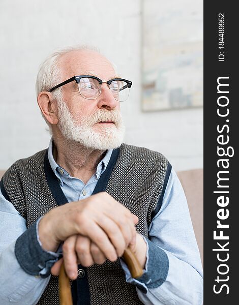 Calm and sad senior man with walking stick looking away, sitting in bright room