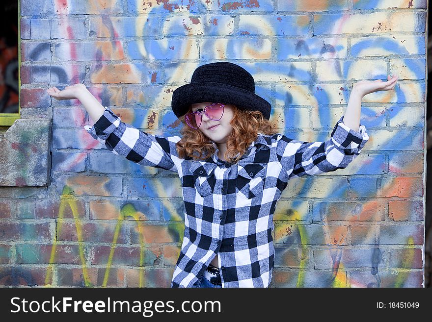 Cute little redheaded girl in black hatand pink glasses with arm up and showing expression in front of brightly painted brick wall. Cute little redheaded girl in black hatand pink glasses with arm up and showing expression in front of brightly painted brick wall