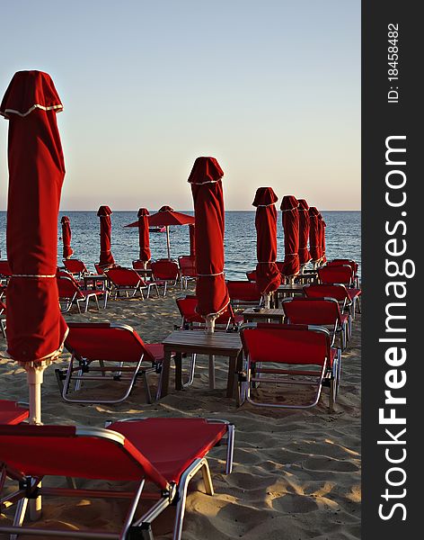 Red sunshades and sunbeds at sandy beach at summer sundown with blue sea and sky in background.