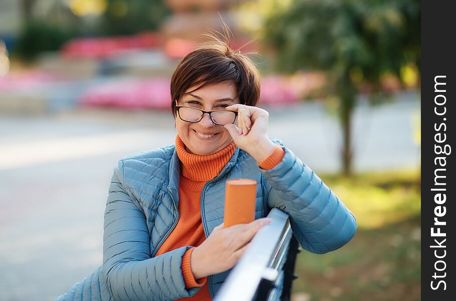 Woman wearing an orange sweater, black glasses sitting on the bench  in the park and smiling
