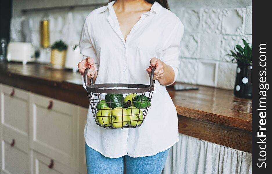 Crop photo of young woman in white shirt hold basket with green fruits and vegetables in hands on the kitchen