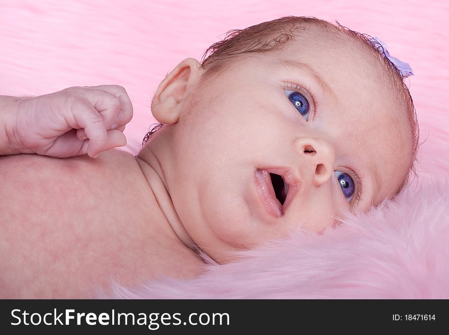 An Adorable Baby With A Pink Background. The baby is wearing purple and has a purple bow in her hair.