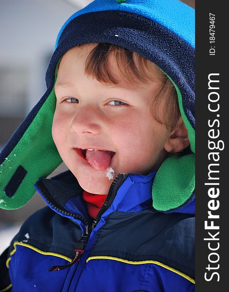A toddler boy dressed in blue and green hat and coat outside playing in the snow and licking a snowflake off his chin. A toddler boy dressed in blue and green hat and coat outside playing in the snow and licking a snowflake off his chin.