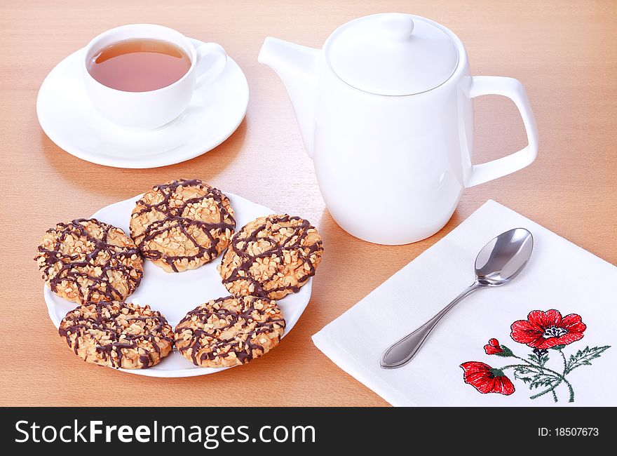 Biscuits with chocolate and peanut decoration on a white plate, teapot and a cup of tea on a brown table background