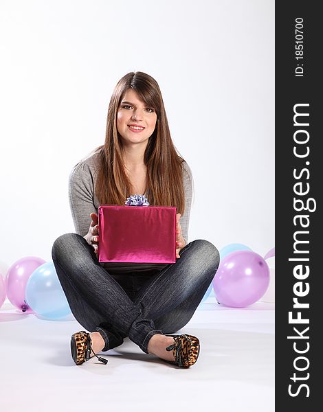 Beautiful young caucasian, student girl with long hair, sitting on the floor with a birthday present. She is wearing a grey top and blue jeans, there are balloons in the background. Beautiful young caucasian, student girl with long hair, sitting on the floor with a birthday present. She is wearing a grey top and blue jeans, there are balloons in the background.