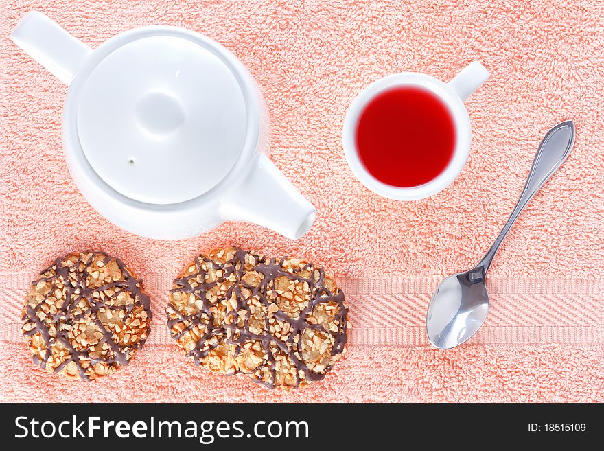 Biscuits with chocolate and peanut decoration on a white plate, teapot and a cup of red tea on a pink napkin background