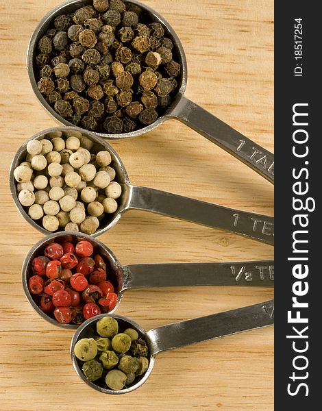 Metal measuring spoons on wood background, each spoon containing different colored whole peppercorns including black, white, red and green. Metal measuring spoons on wood background, each spoon containing different colored whole peppercorns including black, white, red and green