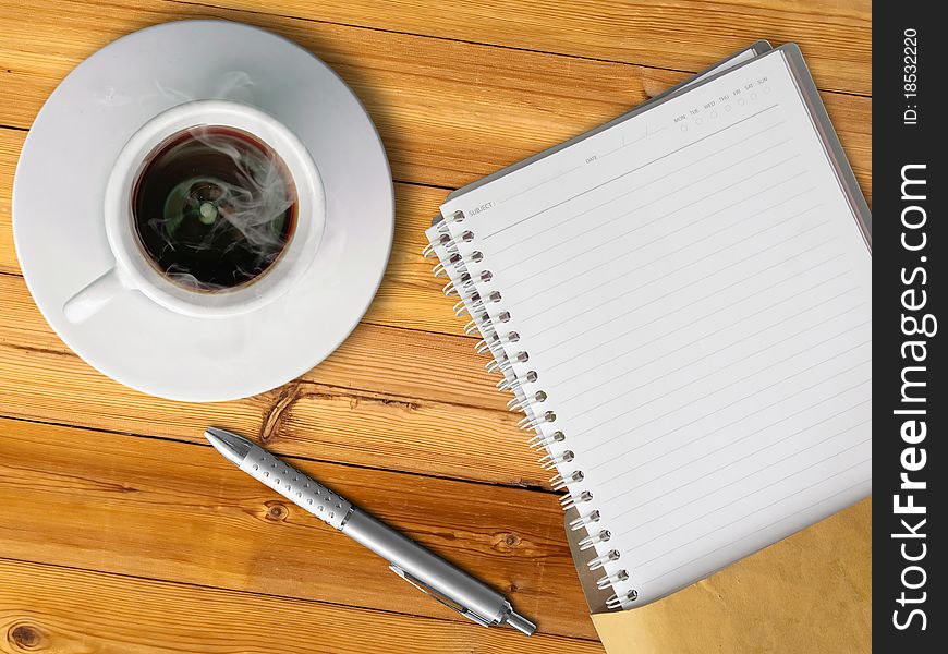 White cup of hot coffee and blank page note book on wood table with silver pen