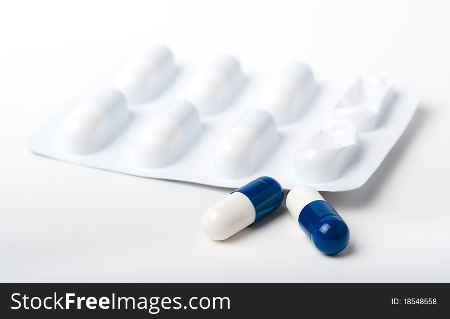 Two blue and white pharmaceutical capsules in front of blister packaging all close up on white. Two blue and white pharmaceutical capsules in front of blister packaging all close up on white.