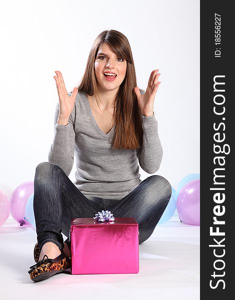 Beautiful young caucasian girl with long hair, has hands raised in happy surprise as she receives a birthday present. Model is wearing jeans and a grey top, sitting on the floor with balloons nearby. Beautiful young caucasian girl with long hair, has hands raised in happy surprise as she receives a birthday present. Model is wearing jeans and a grey top, sitting on the floor with balloons nearby.