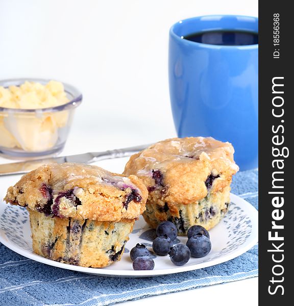 Two blueberry muffins with blueberries on a plate and cup of coffee