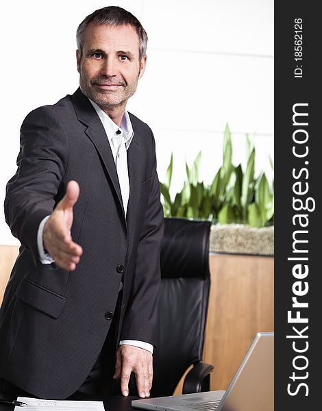 Friendly businessman standing in office behind desk and stretching out hand for handshake, closing a business deal or agreement. Friendly businessman standing in office behind desk and stretching out hand for handshake, closing a business deal or agreement.