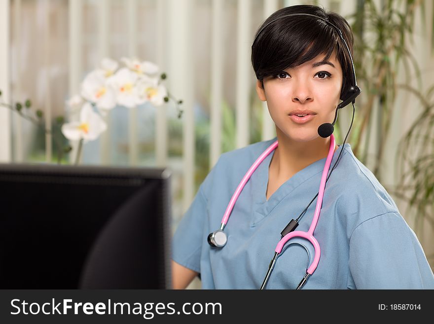 Smiling Attractive Multi-ethnic Young Woman Wearing Headset, Scrubs and Stethoscope Near Her Computer Monitor. Smiling Attractive Multi-ethnic Young Woman Wearing Headset, Scrubs and Stethoscope Near Her Computer Monitor.