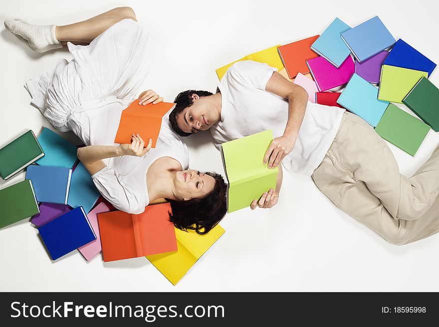 Smiling young man and woman lying on floor with colorful books and reading, looking at books, isolated on white background. Smiling young man and woman lying on floor with colorful books and reading, looking at books, isolated on white background.