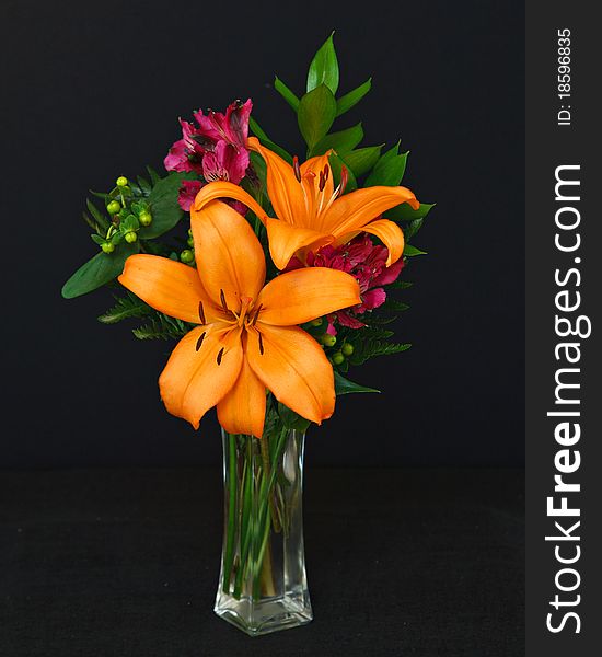 A vibrant bouquet of orange lilies, magenta freesias and greenery in a glass vase, on a black background. A vibrant bouquet of orange lilies, magenta freesias and greenery in a glass vase, on a black background