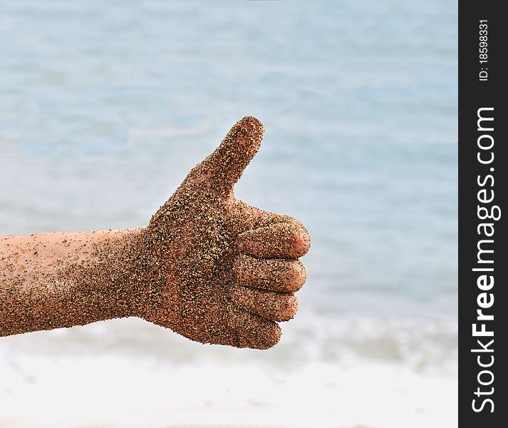 Beach fun with a wet hand with sand on it giving a thumbs up. Ocean beach waves in the background.